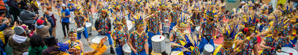 Comparsa playing drums in the street wearing blue and yellow headpieces, multicolor shirts and bllue pants, surrounded by crowd